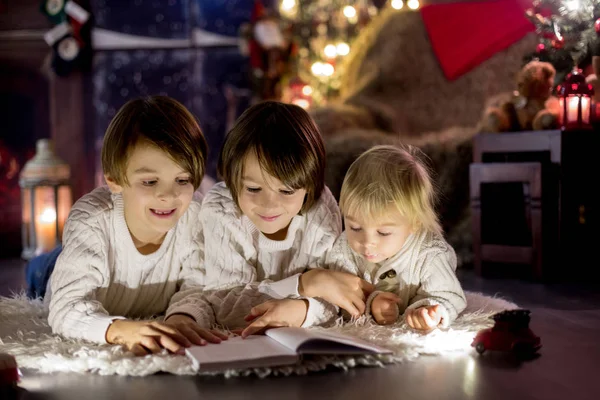 Three children, boy brothers, reading book at home at night on C — Stockfoto