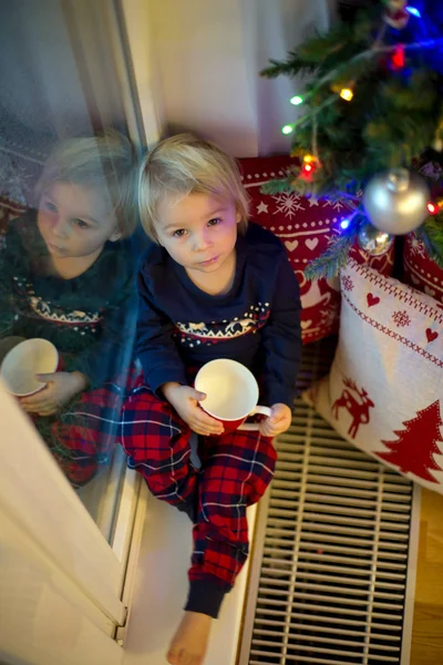 Söt liten pojke i jul pyjamas, sitter på fönstret nära t — Stockfoto