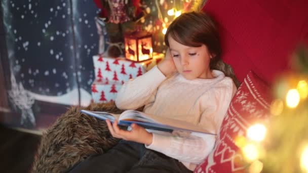 Aranyos fiú, karácsonyfa körül ül egy kényelmes székben, könyvet olvas karácsony este