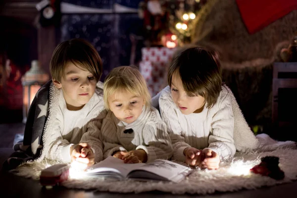 Tres niños, hermanos, leyendo libro en casa por la noche en C —  Fotos de Stock