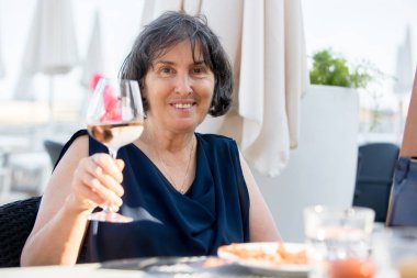 Yaşlı kadın, sahilde bir bardak gül şarabı içiyor 