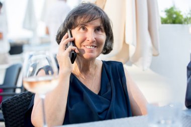 Yaşlı bir kadın, sahilde açık hava restoranında oturuyor, konuşuyor.