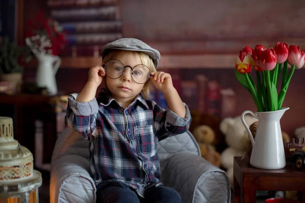 Menino doce, usando óculos, lendo um livro e bebendo — Fotografia de Stock