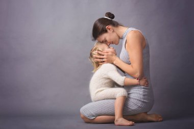 Genç anne, güzel sarışın bebeğine sarılıyor, gri arka planda soyutlanmış.