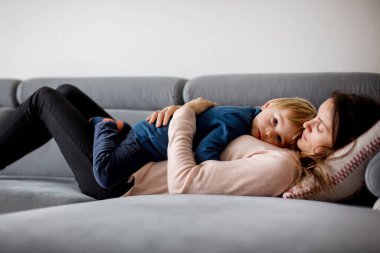 Anne hasta bir çocuğu kucağında tutuyor, ateşli bir şekilde kanepede uzanıyor, burnu ve ateşi akıyor, dinleniyor.