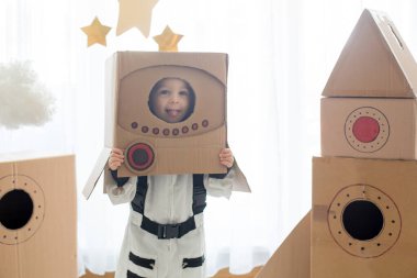 Tatlı çocuk, astronot gibi giyinmiş, evde karton roket ve kutudan yapılmış el yapımı kaskla oynuyor.