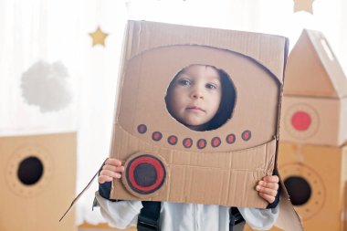Tatlı çocuk, astronot gibi giyinmiş, evde karton roket ve kutudan yapılmış el yapımı kaskla oynuyor.