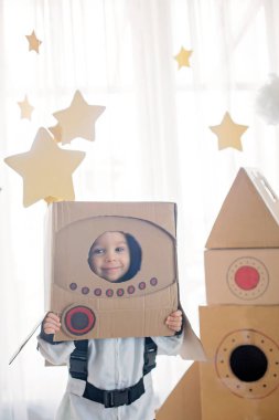 Tatlı çocuk, astronot gibi giyinmiş, evde karton roket ve kutudan yapılmış el yapımı kaskla oynuyor.