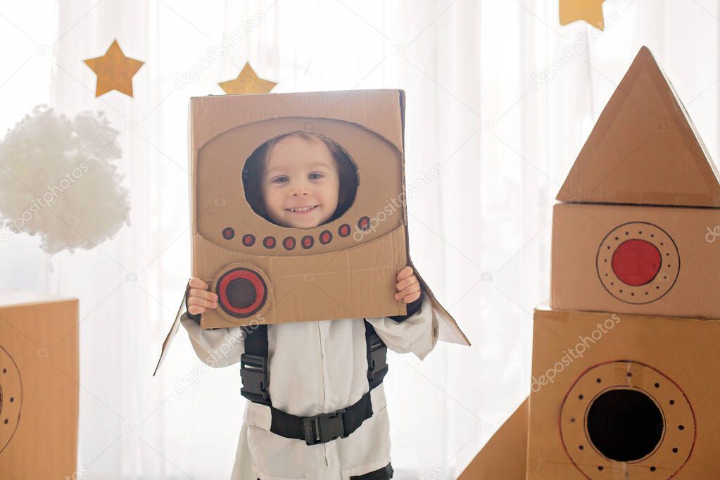 Sweet toddler boy, dressed as an astronaut, playing at home with cardboard rocket and handmade helmet from box