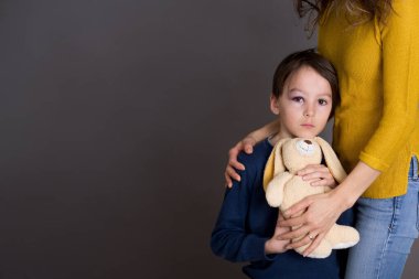 Gözü morarmış üzgün bir çocuk, yaralı bir göz, annesine ve oyuncak ayısına sarılmış.