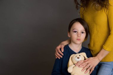 Gözü morarmış üzgün bir çocuk, yaralı bir göz, annesine ve oyuncak ayısına sarılmış.
