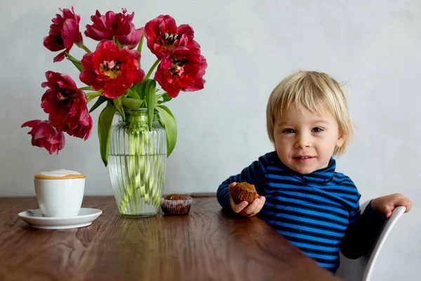 小さな幼児ブロンドの男の子 リビングルームで自宅でチョコレートマフィンを食べる テーブルの上に花と美しい花瓶 — ストック写真