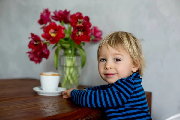 小さな幼児ブロンドの男の子 リビングルームで自宅でチョコレートマフィンを食べる テーブルの上に花と美しい花瓶 — ストック写真