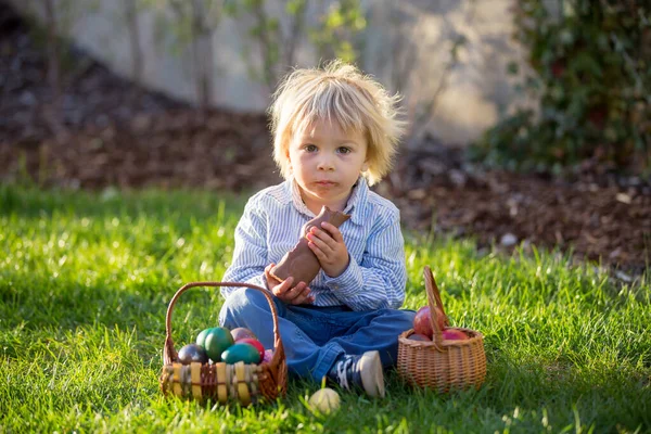 蹒跚学步的小男孩 日落时在花园里吃着巧克力兔子 身边是复活节彩蛋 — 图库照片
