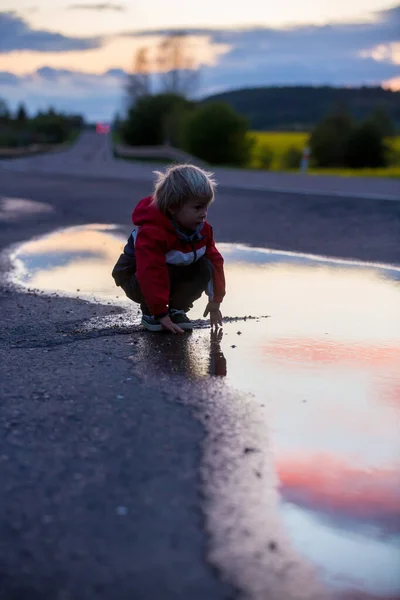 甘い幼児の子供 赤いレインコートのかわいい男の子 日没の水たまりで遊ぶ — ストック写真