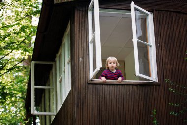 Sevimli çocuk, ormandaki dağ evinin penceresinden dışarı bakıyor, bahar zamanı.