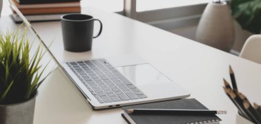 Dizüstü bilgisayar ve ofis malzemeleriyle pencerelerin yanındaki beyaz masada rahat çalışma alanının görüntüsü.