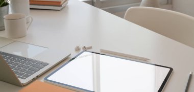 Tablet, dizüstü bilgisayar ve ofis malzemeleriyle pencerelerin yanındaki beyaz masada rahat çalışma alanının görüntüsü.