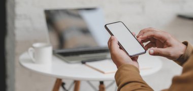 Arka planda dizüstü bilgisayarı ve ofis malzemeleri olan boş ekran akıllı telefonu tutan genç adamın yakın görüntüsü 
