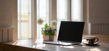 Laptop bilgisayarı, kahve fincanı ve pencerelerin yanındaki ahşap masa üzerinde rahat bir iş yeri. 