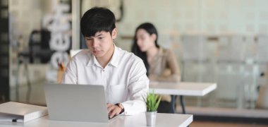 Genç profesyonel erkek tasarımcı arka planda iş arkadaşıyla dizüstü bilgisayar kullanırken projesinde çalışıyor.