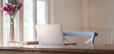 Dizüstü bilgisayar ve ofis malzemeleriyle pencerelerin yanındaki ahşap masada rahat bir iş yeri görüntüsü.