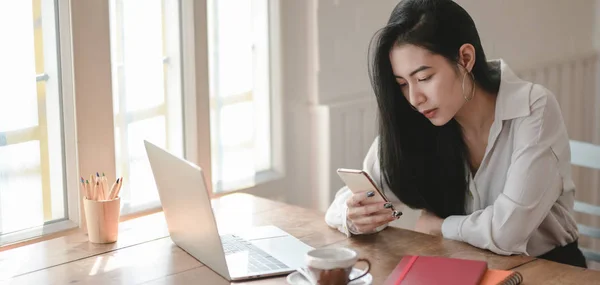 Porträt einer schönen Geschäftsfrau, die an ihrem Projekt arbeitet, während sie ihr Smartphone am bequemen Arbeitsplatz benutzt — Stockfoto