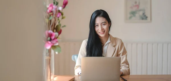 Porträt einer jungen Geschäftsfrau, die mit Laptop an ihrem Projekt arbeitet, während sie in die Kamera lächelt — Stockfoto