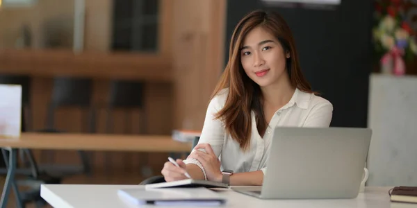 Молода красива азіатська бізнес-леді працює над своїм проектом з ноутбуком і посміхається до камери — стокове фото