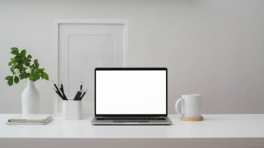 Boş ekran dizüstü bilgisayar, çerçeve, kalemler, kahve fincanı ve vazo ile beyaz duvarlı beyaz masa 