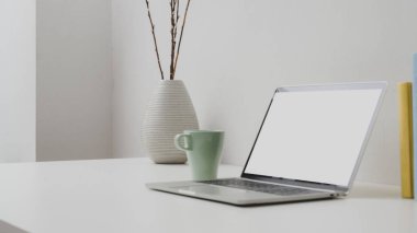 Boş ekran laptop, kahve fincanı ve beyaz duvarlı seramik vazo ile çalışma alanının kırpılmış görüntüsü. 