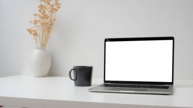 Boş ekran laptop, kahve fincanı ve beyaz duvarlı seramik vazo ile çalışma alanının kırpılmış görüntüsü. 