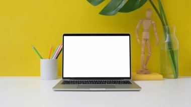 Boş ekran dizüstü bilgisayarı, kalemler, ahşap figür ve sarı arka planlı beyaz masa üzerindeki vazo ile kırpılmış çalışma alanı. 