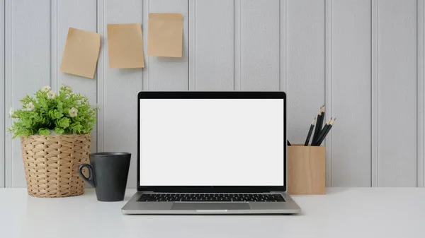 Vista ravvicinata dell'area di lavoro con computer portatile con schermo bianco, cancelleria, nota adesiva, tazza di caffè e vaso di albero su tavolo bianco con parete a listello — Foto Stock