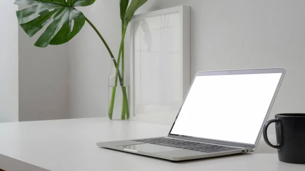 Закрыть вид на рабочее пространство ноутбуком с экраном Blank, чашкой кофе, рамой и вазой на столе с белой стеной — стоковое фото