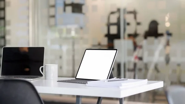 Imagen recortada del lugar de trabajo con tableta de pantalla en blanco, computadora portátil, suministros de oficina y taza de café en la mesa blanca con sala de oficina borrosa —  Fotos de Stock