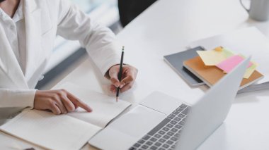 Ofis odasındaki ofis malzemeleriyle dizüstü bilgisayarda çalışırken iş kadınının not defterine yazdığı kısa bir kare.