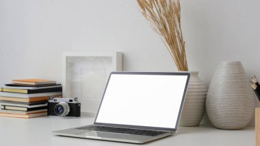 Boş ekran dizüstü bilgisayar, kamera, çerçeve, kitap ve dekorasyonla beyaz masa üzerinde beyaz duvar arkaplanlı kesilmiş iş yeri görüntüsü
