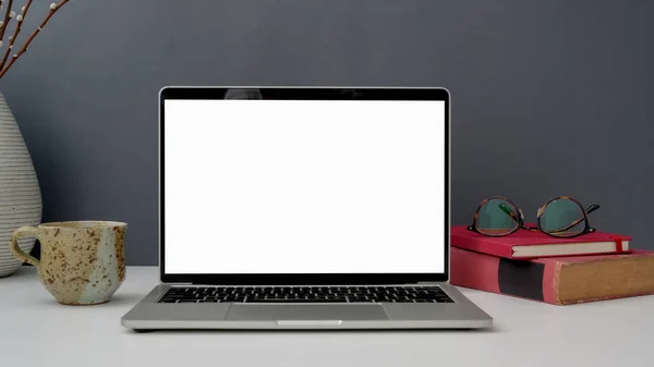 Vista ravvicinata dell'area di lavoro con computer portatile, tazza, occhiali e libri aperti su tavolo bianco con sfondo grigio a parete — Foto Stock