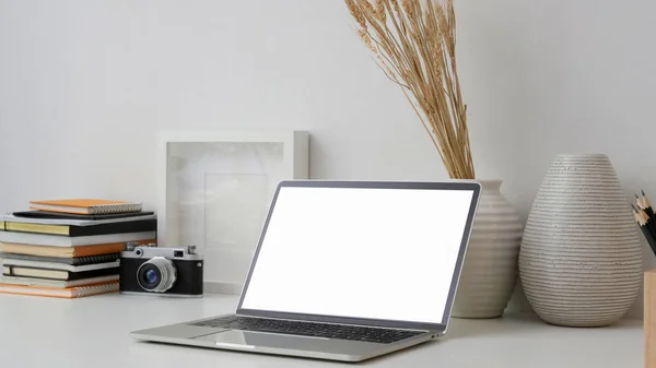 Recorte de local de trabalho com tela em branco laptop, câmera, quadro, livros e decorações na mesa branca com fundo de parede branca — Fotografia de Stock