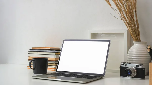 Recorte de local de trabalho com tela em branco laptop, câmera, quadro, livros e decorações na mesa branca com fundo de parede branca — Fotografia de Stock