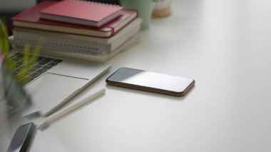 Beyaz masa arkasındaki boş ekran akıllı telefonu, dizüstü bilgisayar, kopyalama alanı ve ofis malzemelerinin yan görünümü