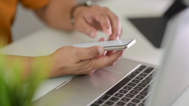 Basit bir ofis odasında dizüstü bilgisayarla çalışırken akıllı telefondan yazı yazan serbest çalışan erkek.