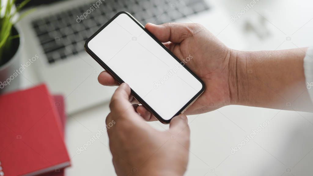 Cropped shot of businessman holding smartphone in workspace with other office supplies on blurred white table background