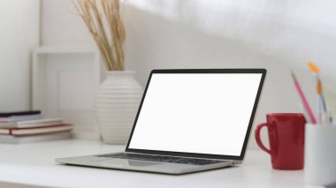 Boş ekran dizüstü bilgisayarı, ofis malzemeleri, boya fırçası ve beyaz masa süslemeleri olan şık bir ofis odası. 