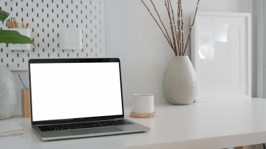 Boş ekran laptop ile modern çalışma alanının kırpılmış görüntüsü, beyaz masa üzerinde çerçeve ve seramik vazo 