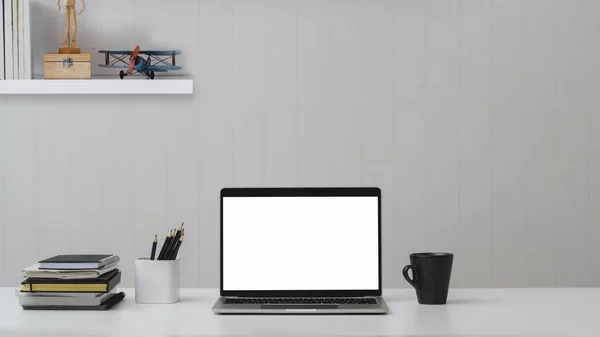 Vista cercana del lugar de trabajo con computadora portátil de pantalla en blanco, suministros de oficina y taza de café en la mesa blanca con estante en la pared blanca —  Fotos de Stock