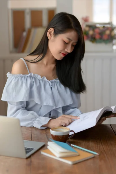 Nahaufnahme eines Mädchens, das Buch liest, während es in einem komfortablen Zimmer mit Laptop und Schreibwaren sitzt — Stockfoto