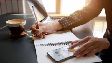 Side view of female writing on notebook while looking business chart on smartphone with laptop and other office supplies in comfortable workplace