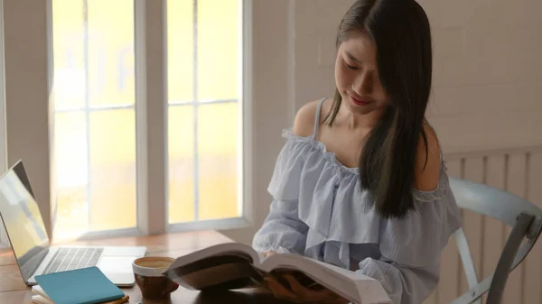 Recorte tiro de um livro de leitura menina enquanto se situa ao lado da janela em quarto confortável com laptop e artigos de papelaria — Fotografia de Stock